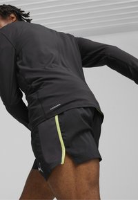 Schwarzes, sportliches Langarmshirt und Shorts mit neongrünen Akzenten. Das Shirt hat ein tailliertes Design; die Shorts verfügen über Mesh-Seitenpaneele und ein Logo.