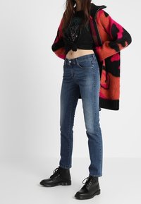 Pletený kardigan v růžové a černé barvě s odvážnými vzory, černé zkrácené tričko, modré úzké džíny a černé šněrovací kotníkové boty.