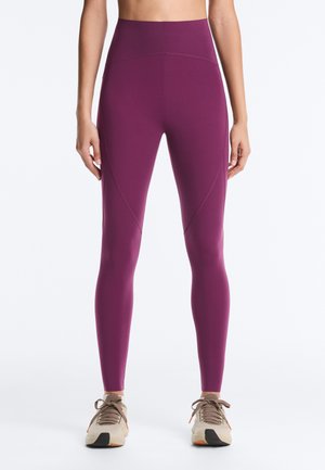 Personne portant des leggings violets taille haute et des baskets beiges, debout avec les bras détendus le long du corps sur fond blanc.