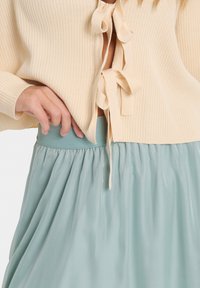 Cardigan beige lavorato a maglia con lacci frontali, abbinato a una gonna in satin azzurro chiaro con vita elastica e texture arricciata.