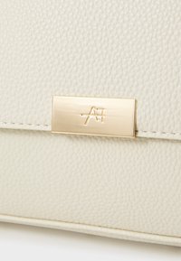 Sac à main en cuir beige clair avec une surface texturée, présentant une plaque logo en métal doré et un fermoir lisse.