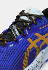 Μωβ και μπλε αθλητικό παπούτσι με υφασμάτινο επάνω μέρος, μαύρα και πορτοκαλί κορδόνια, και ένα εμφανές χρυσό λογότυπο. Διαθέτει ανθεκτική σόλα και ενισχυμένη μύτη.