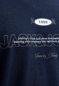 Tamsiai mėlynas medvilninis marškinėlis su ekranu spaustu tekstu: "JACK&JONES," "1990," ir šūkiu balta ir šviesiai pilka spalvomis. Minkšta tekstūra.