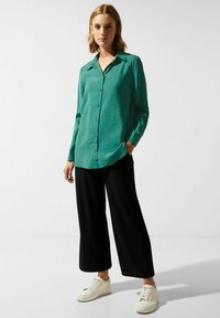 Green button-up shirt with a pointed collar, long sleeves, and a relaxed fit. Paired with black wide-leg pants and white sneakers.