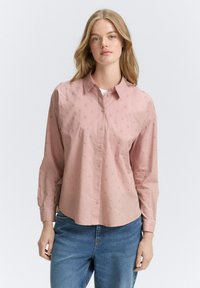Camicia button-up rosa chiaro con una texture morbida, caratterizzata da un piccolo motivo a pois e maniche lunghe. Abbinata a jeans blu.