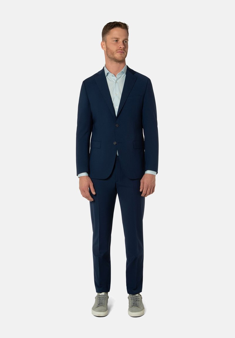 Marineblauer maßgeschneiderter Anzug mit einem Zwei-Knopf-Jacket und einer slim-fit Hose. Darunter ein hellblaues Hemd, kombiniert mit grauen Freizeit-Sneakern.