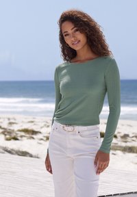 Woman with curly hair wearing green long-sleeve top and white pants standing on boardwalk by the beach with ocean background.