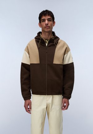 Mand iført en color-blocked hættetrøje i brun, beige og creme med cremefarvede bukser mod en ensfarvet lys baggrund.