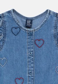 Džinsinis drabužis su apvalia iškirpte, mėlynais mygtukais ir raudonos, tamsiai mėlynos bei baltos spalvų širdies formos siuvinėjimu. Su raukšlėtu peties dekoravimu.