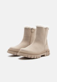 Beige nilkkurit, joissa on pehmeä, teksturoitu keinoturkisreuna, vetoketjukiinnitys sivulla ja paksu, uritettu pohja pitämään otetta.