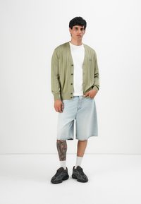 Homem vestindo um cardigan verde claro abotoado, camiseta branca, calções de denim claros até ao joelho, meias brancas e ténis pretos, em pé com uma mão no bolso.