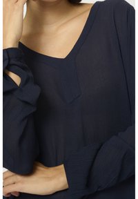 Marineblå bluse med V-hals design, tekstureret stof og lange ærmer med rynkede detaljer nær manchetterne.