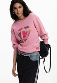 Rožinis džemperis su širdies dizainu ir įvairiaspalviais raštais. Yra riboti rankogaliai ir etiketė apačioje. Derinamas su mišinio džinsais ir juoda rankine.