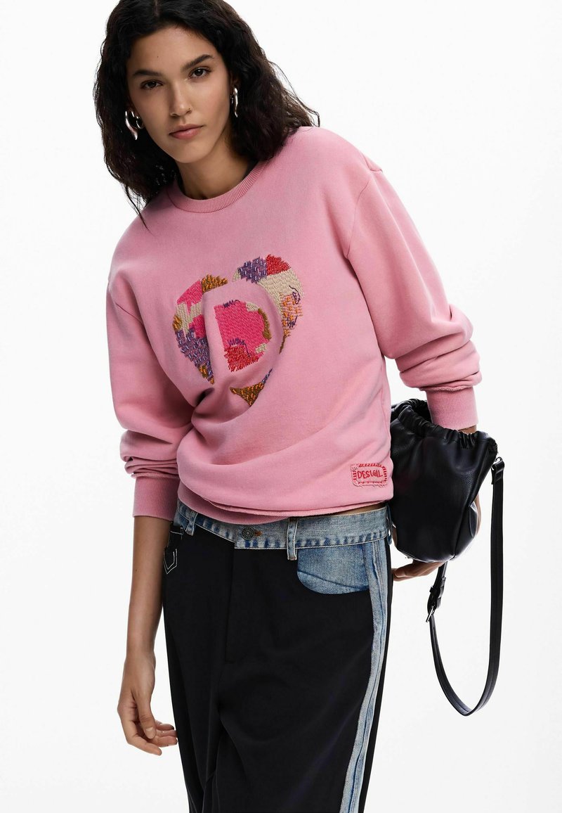 Rožinis džemperis su širdies dizainu ir įvairiaspalviais raštais. Yra riboti rankogaliai ir etiketė apačioje. Derinamas su mišinio džinsais ir juoda rankine.