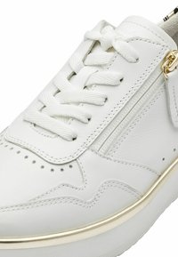 White leather sneaker featuring intricate perforations, flat white laces, side zippers, and a gold accent on the heel. Textured midsole.