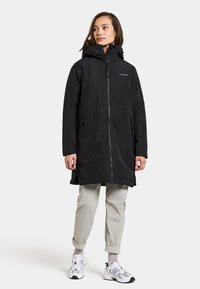 Parka nera impermeabile con cappuccio, zip frontale e tasche laterali. Presenta polsini elasticizzati e una vestibilità rilassata, indossata sopra pantaloni grigi chiari.