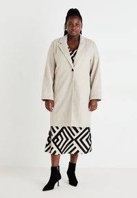 Beige uldfrakke med klapkrave og single knaplukning, båret over en mønstret sort og beige nederdel. Sorte ankelstøvler fuldender outfittet.
