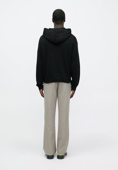 Persoană îmbrăcată cu un pulover cu glugă negru, pantaloni largi bej și încălțăminte neagră, stând cu spatele împotriva unui fundal alb simplu.