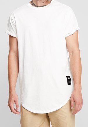 Biała koszulka bawełniana z podwiniętymi rękawami i zaokrąglonym dołem. Ma małą czarną metkę z nadrukowanym tekstem po boku. Gładka tekstura.