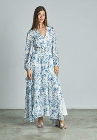 Maxikleid aus leichtem, transparentem Stoff mit blauem Blumendruck, langen Ärmeln, tiefem V-Ausschnitt, rüschigem Saum und passendem Gürtel. Silbern hohe Sandalen.