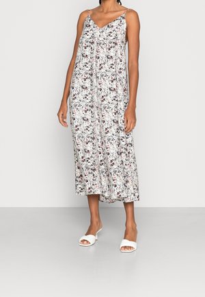 Femme portant une robe midi blanche sans manches avec fines bretelles et un petit imprimé floral, associée à des sandales blanches à talons ouverts.