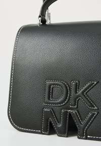 Melna ādas somiņa ar teksturētu virsmu, ar izteiktu DKNY logo paceltās burtu formās un baltu šuvumu gar malām.