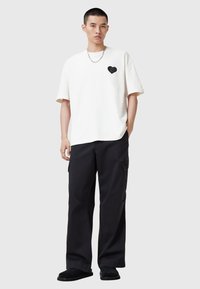 Camiseta de algodón blanca con gráfico de corazón negro en el pecho, combinada con pantalones cargo negros. El modelo lleva sandalias deslizantes negras y un collar de cadena plateado.