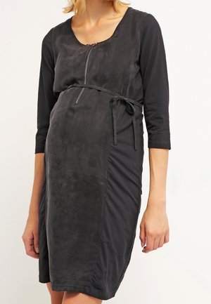 Kobieta w czarnej sukience o długości do kolan, z rękawami do łokcia, zamkiem błyskawicznym z przodu i paskiem związanym w talii.