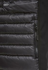 Svart puffer vest med kviltet tekstur, synlig glidelås og tøymerke; kombinerer skinnende og matte overflater.