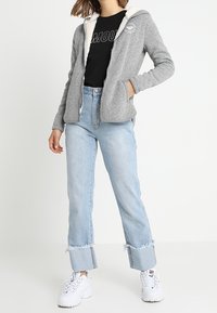 Felpa grigia con zip e foderata in pile bianco, abbinata a una maglietta nera e jeans high-waisted azzurri chiari con orli sfrangiati e sneaker bianche.