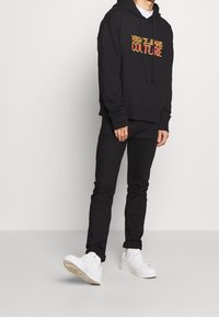 Svart hoodie med en röd och gul "Versace Jeans Couture" logga, kombinerad med svarta byxor och vita sneakers. Avslappnad streetwear-stil.
