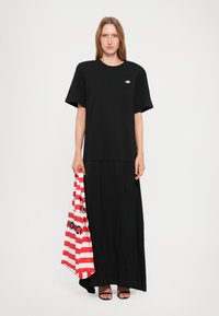 Woman wearing a black oversized T-shirt and long skirt, holding a red and white striped tote bag, standing on a white background.