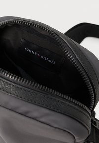 Czarna nylonowa torba typu crossbody z zapięciem na zamek, charakteryzująca się gładką teksturą oraz wewnętrzną metką z logo, prezentująca elegancki design i subtelne detale.