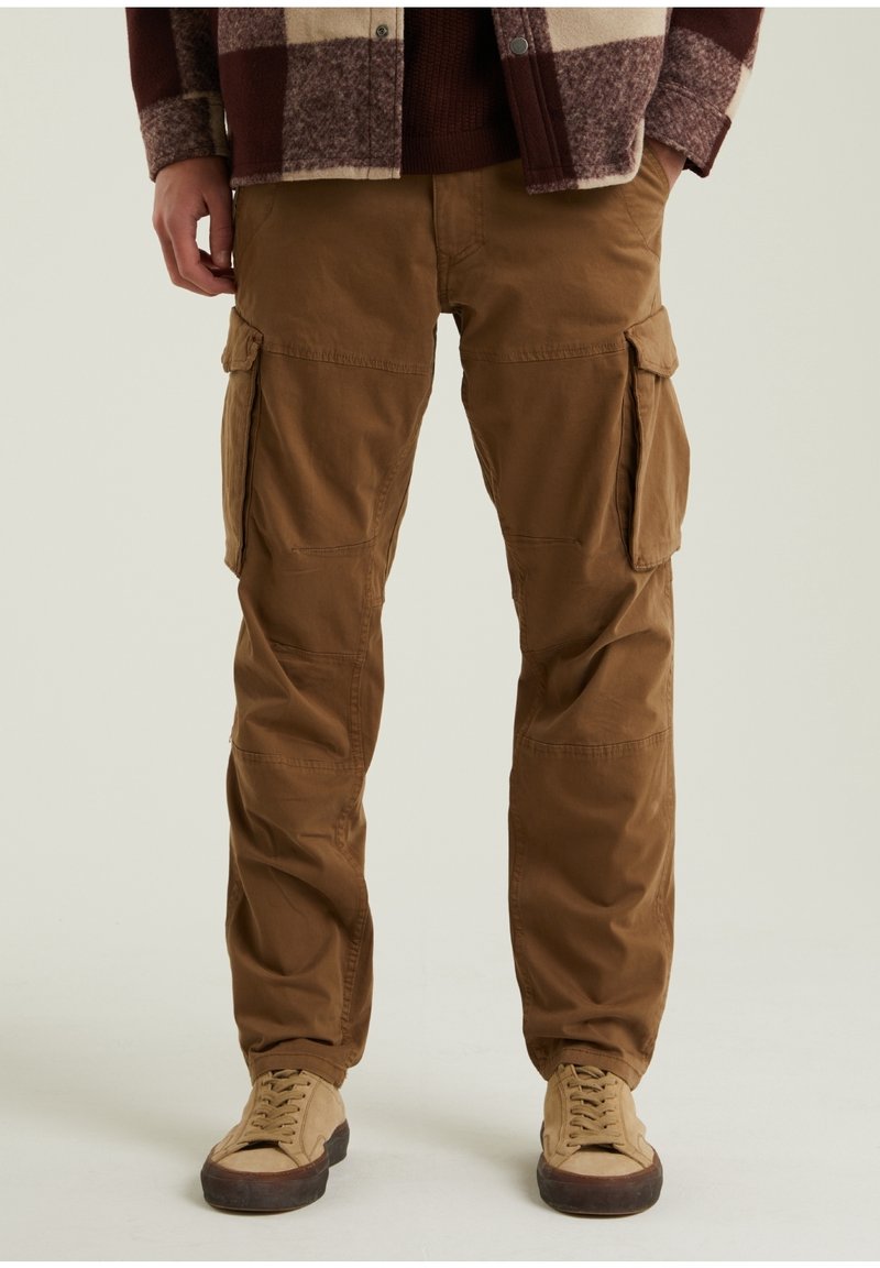 Pantalon cargo marron avec poches latérales porté avec des baskets beiges et une veste à carreaux, visible de la taille jusqu'aux chaussures.