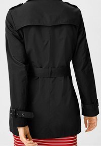 Trench-coat noir avec une taille ceinturée, des manches longues et des épaulettes. Texture lisse, longueur mi-mollet, assorti à une jupe rayée.