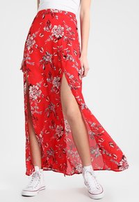 Jupe longue rouge à motifs floraux avec fentes hautes, tissu léger, motif floral blanc et ourlet roulé ; portée avec des baskets blanches pour le contraste.