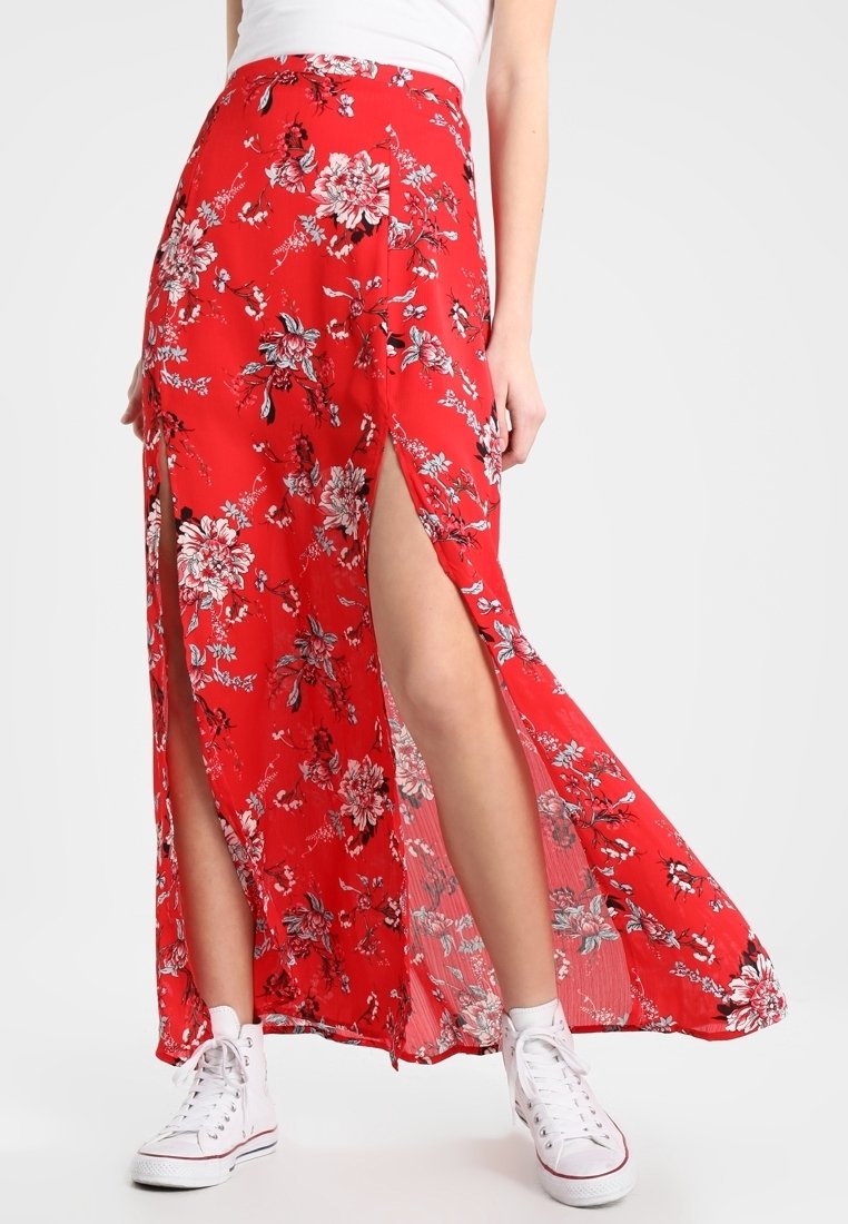 Jupe longue rouge à motifs floraux avec fentes hautes, tissu léger, motif floral blanc et ourlet roulé ; portée avec des baskets blanches pour le contraste.