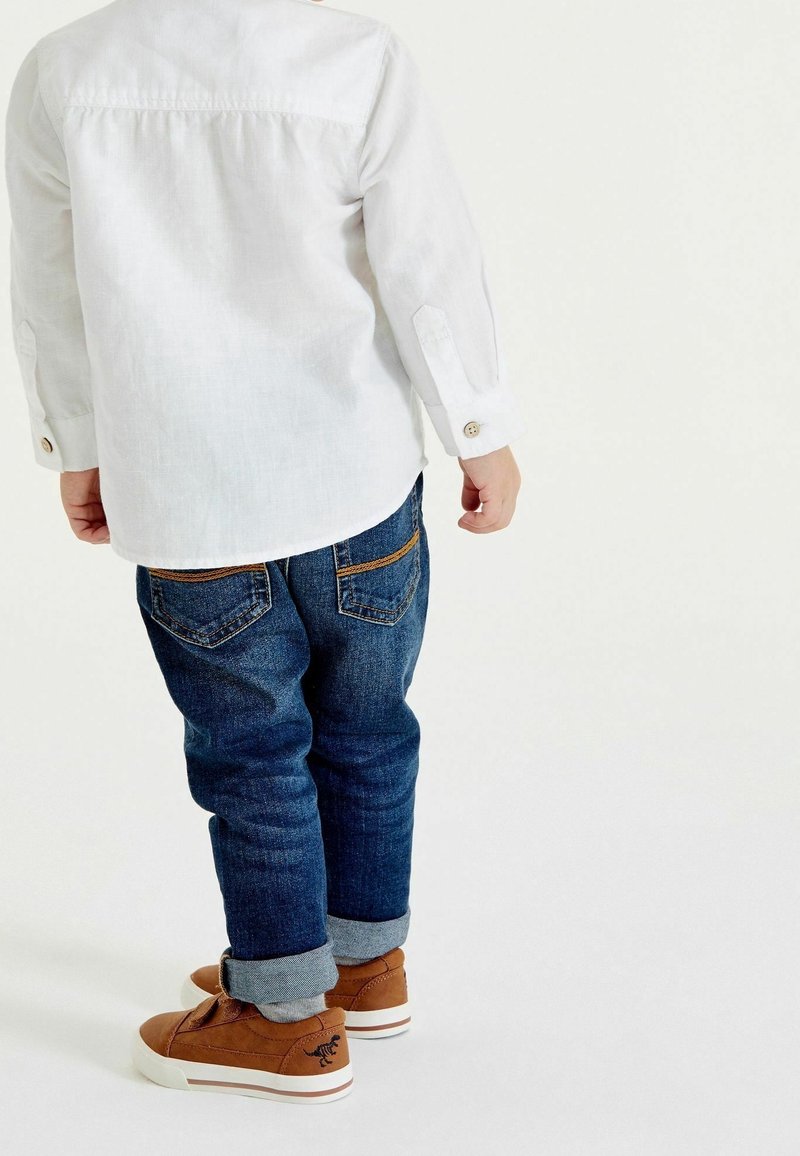 Enfant en t-shirt blanc à manches longues, jean bleu retroussé et baskets marron, debout de dos sur un fond blanc uni.