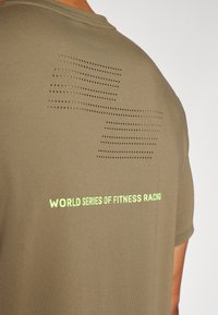 T-shirt atlético verde-oliva com padrão de malha perfurada nas costas e texto "WORLD SERIES OF FITNESS RACING" em amarelo-verde vibrante.