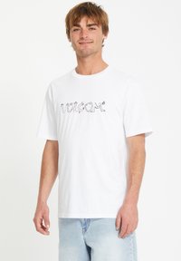 T-shirt en coton blanc avec un logo "Volcom" stylisé noir sur le devant. Manches courtes et coupe décontractée. Assorti avec un jean bleu clair.