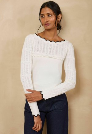 Woman with dark hair in a low bun wears a white long-sleeve top with textured patterns and black scalloped edges, paired with dark jeans.