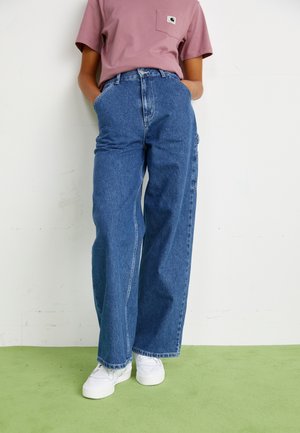 Person trägt hoch taillierte, weit geschnittene blaue Jeans, ein mauvefarbenes kurzärmeliges Hemd mit Brusttasche und weiße Turnschuhe und steht auf grünem Boden.