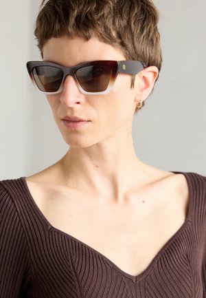 Gafas de sol marrones en forma de ojo de gato con lentes degradados, un grueso marco de carey y patillas oscuras contrastantes que presentan un sutil detalle de logotipo.