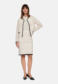 Weiße strukturierte Strickjacke mit schwarzem Besatz, Knopfverschluss und passendem Rock. Das Outfit zeichnet sich durch aufwendige Muster und eine taillierte Silhouette aus.