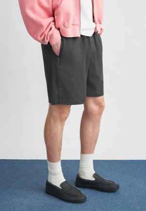 Σκούρα γκρι σορτς με τσέπες, συνδυασμένα με μαύρα παπούτσια slip-on και λευκές κάλτσες. Το μοντέλο φοράει ροζ cropped φούτερ πάνω από ένα λευκό T-shirt.