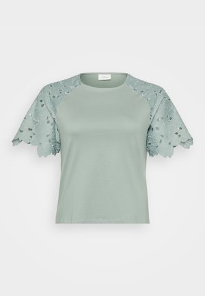 Haut vert clair avec des manches en dentelle présentant des motifs floraux et des détails découpés, mettant en avant un design décontracté mais élégant.