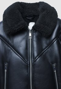 Giacca di pelle nera con ampio colletto in pelliccia sintetica nera e zip frontali e delle tasche in argento, mostrata in primo piano su uno sfondo bianco.