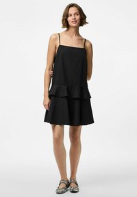 Must värvitu, ilma varrukateta kleit ruudukujulise dekoltee ja kihilise volangiga hemiga, valmistatud siledast kangast. Mudel kannab must-valgeid triibulisi kingi.