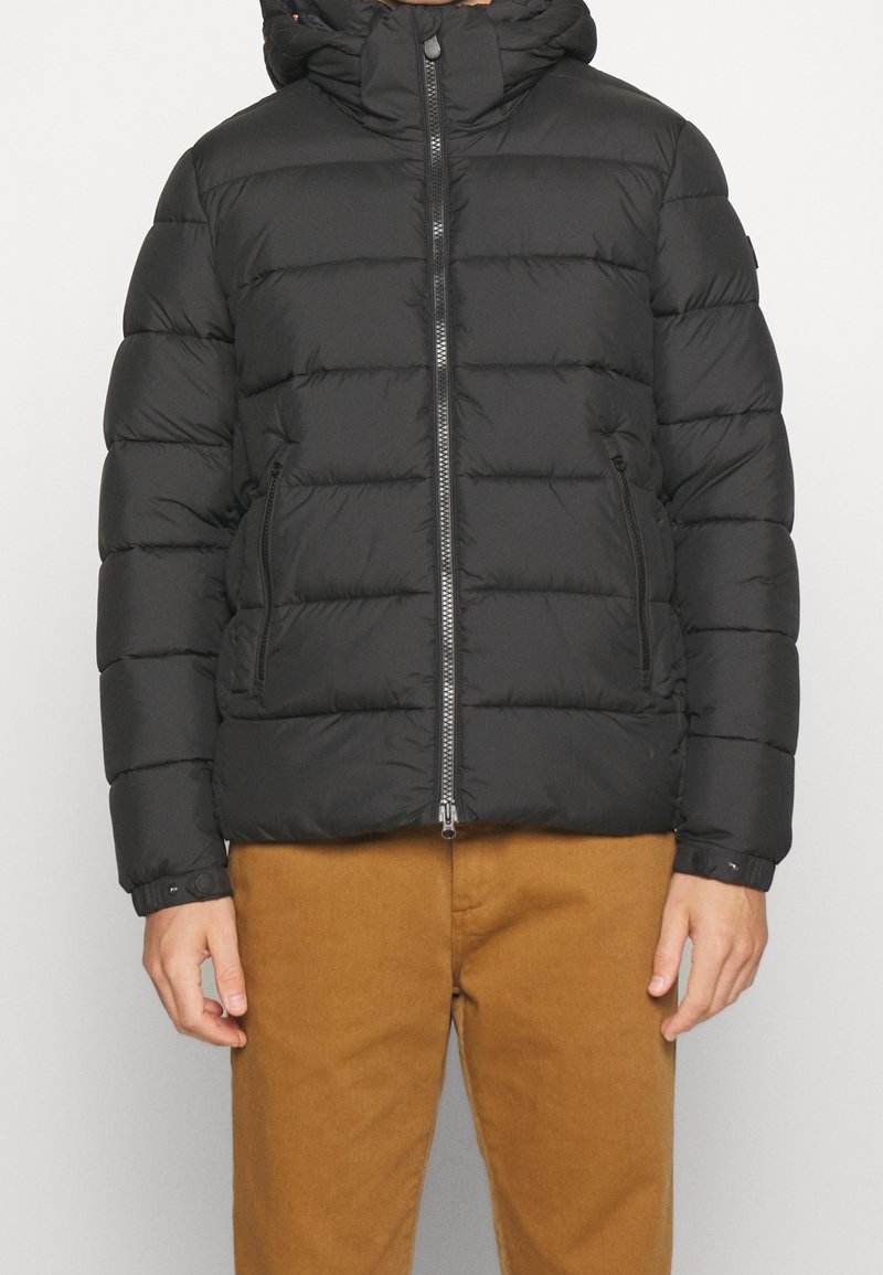 Veste matelassée noire à fermeture éclair avec capuche, deux poches avant zippées et détails de poignets à pression, portée avec un pantalon marron.
