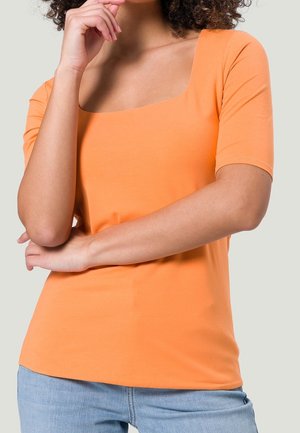 Frau mit lockigem Haar, die ein oranges Kurzarmoberteil mit eckigem Ausschnitt und hellblaue Jeans trägt, während sie nachdenklich ihr Kinn berührt.